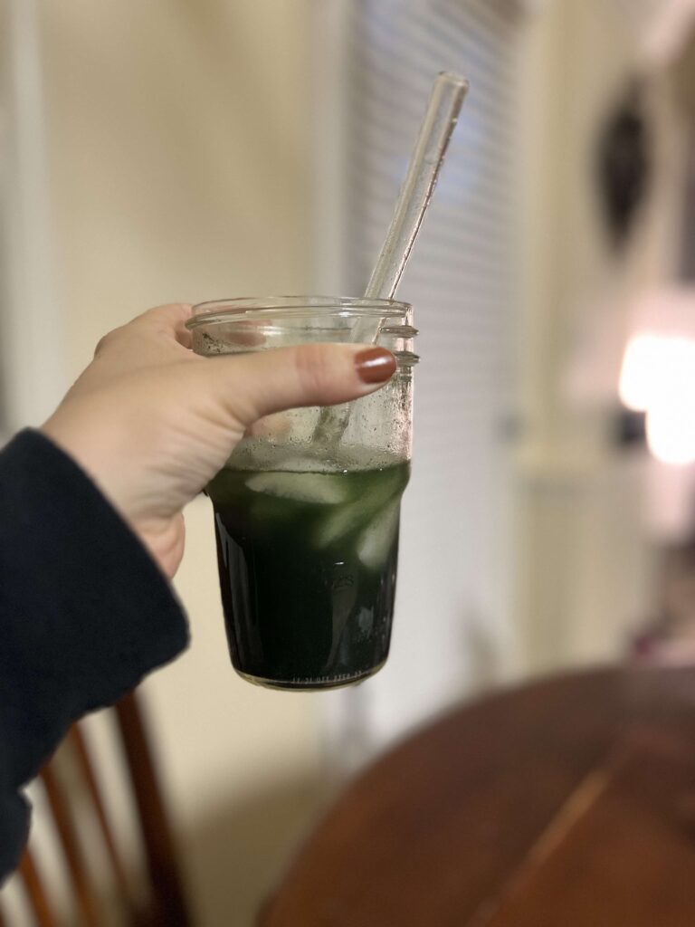 Hand holding an Arbonne GreenSynergy Elixir powder mixed with ice and water and a glass straw